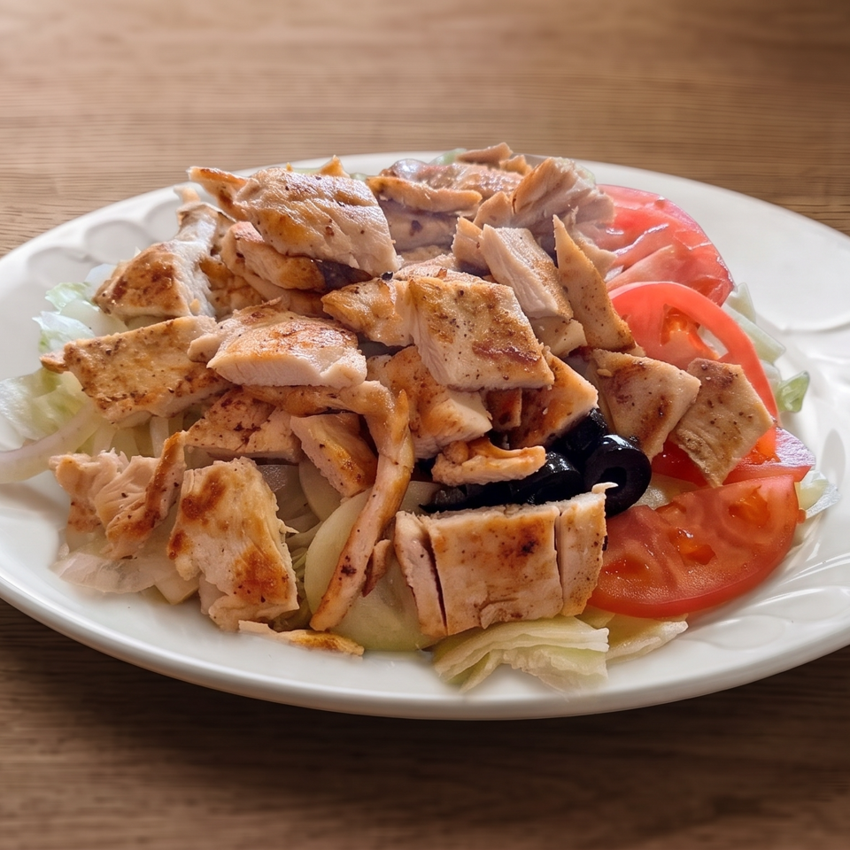 Grilled chicken salad with lettuce, tomatoes, onions, and black olives on a white plate atop a wooden table.